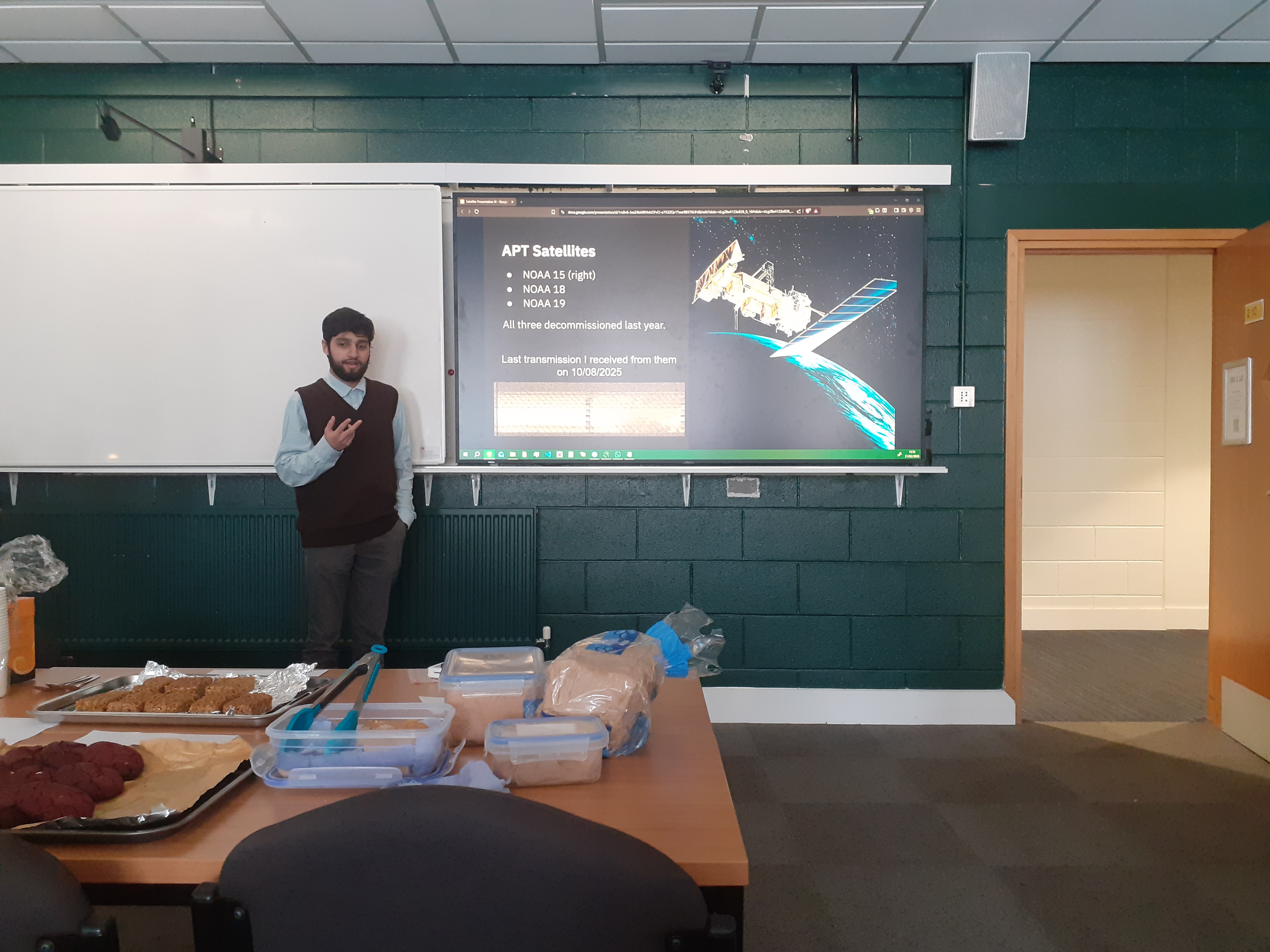 A student is giving a presentation about reciving satellite imagery using amateur radio. There are several cakes for a bake sale infront of the student.
