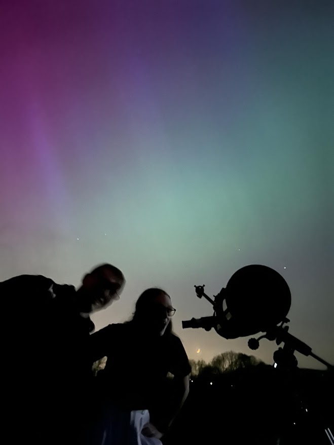 A silhouette of two students and a telescope lie infront of a colourful aurora.