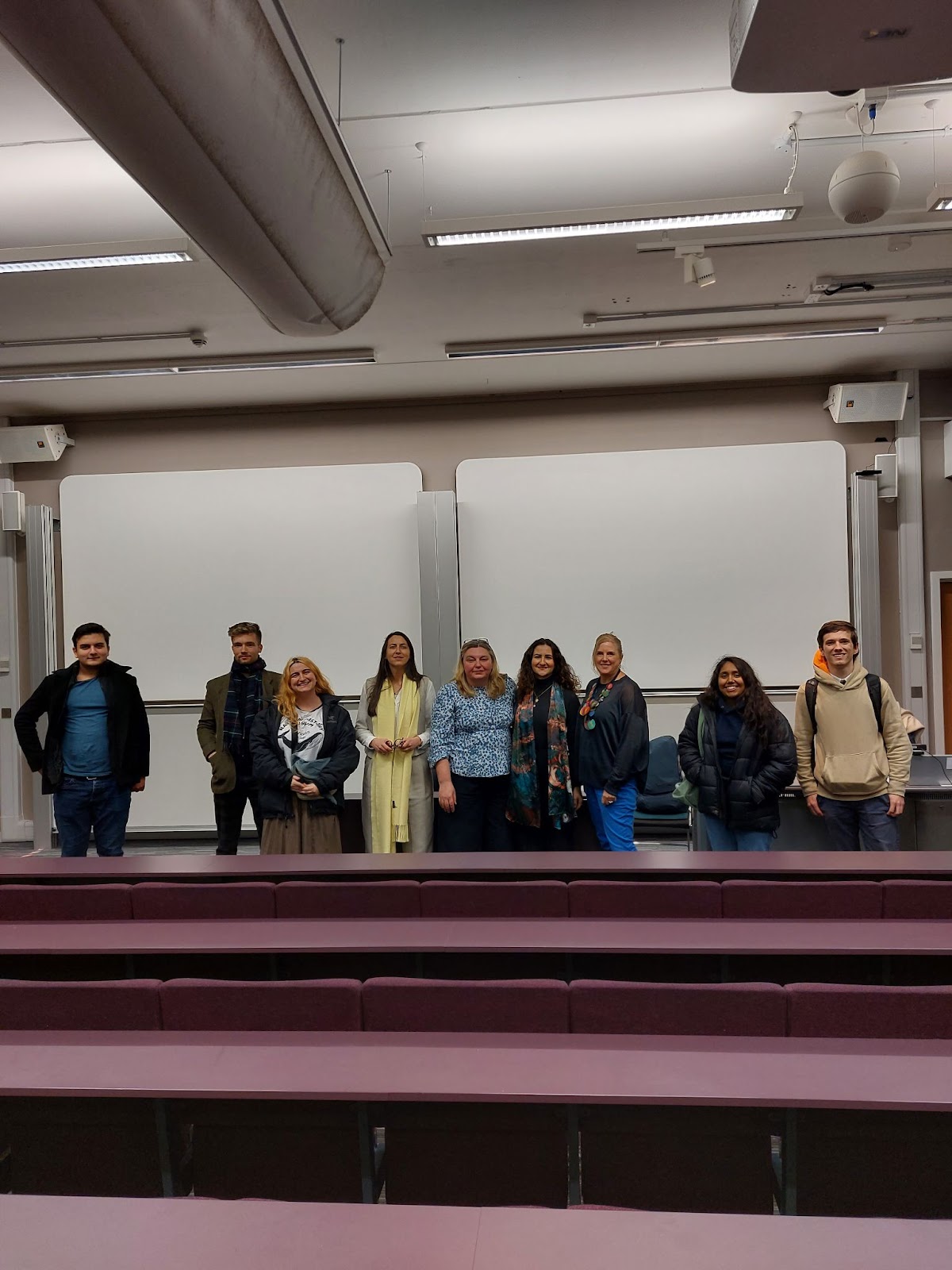 A group of people standing in front of some whiteboards. There are four guest speakers and a number of students