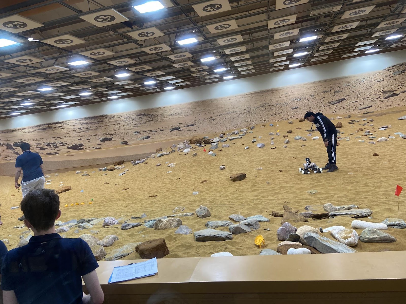 Inside the Mars simulation environment at Airbus. The ground is sandy, with rocks scattered around. There are people standing around controlling rovers, and the ceiling is covered in fluorescent lights.