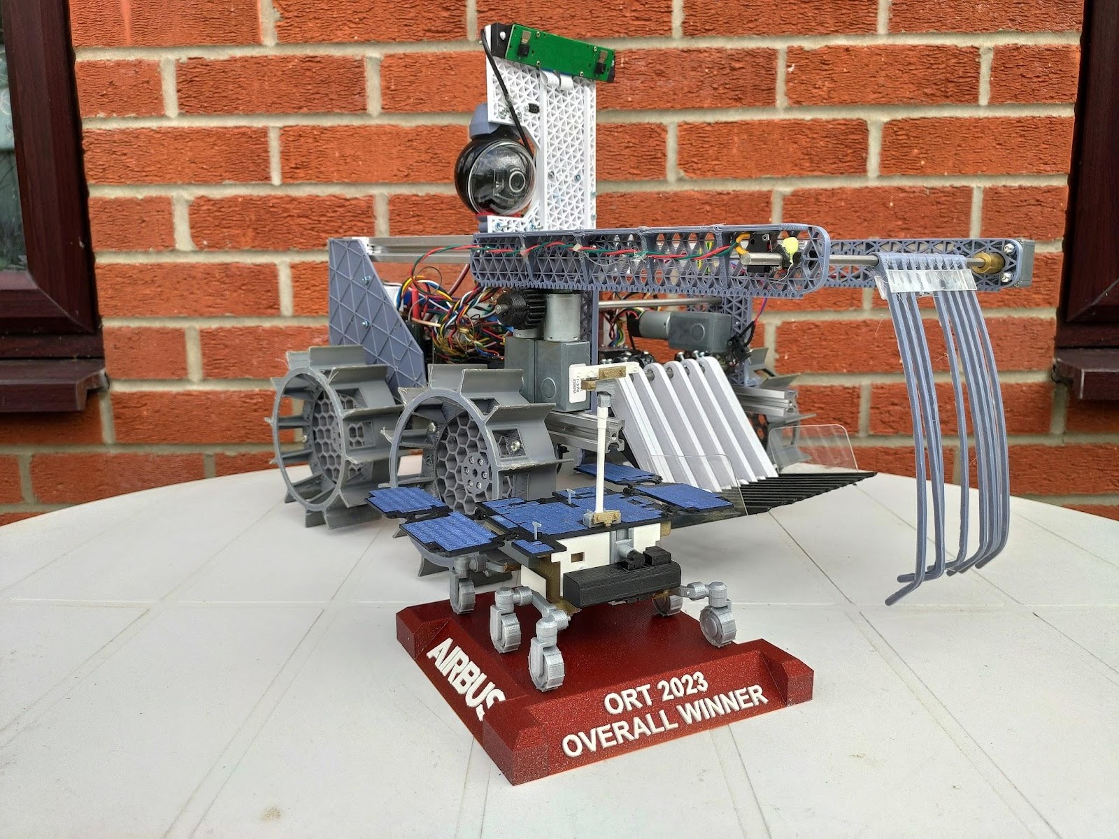 The UKSEDs Roving Trials first place trophy is sitting on a table in front of TBRo's rover. Both are in front of a red brick wall