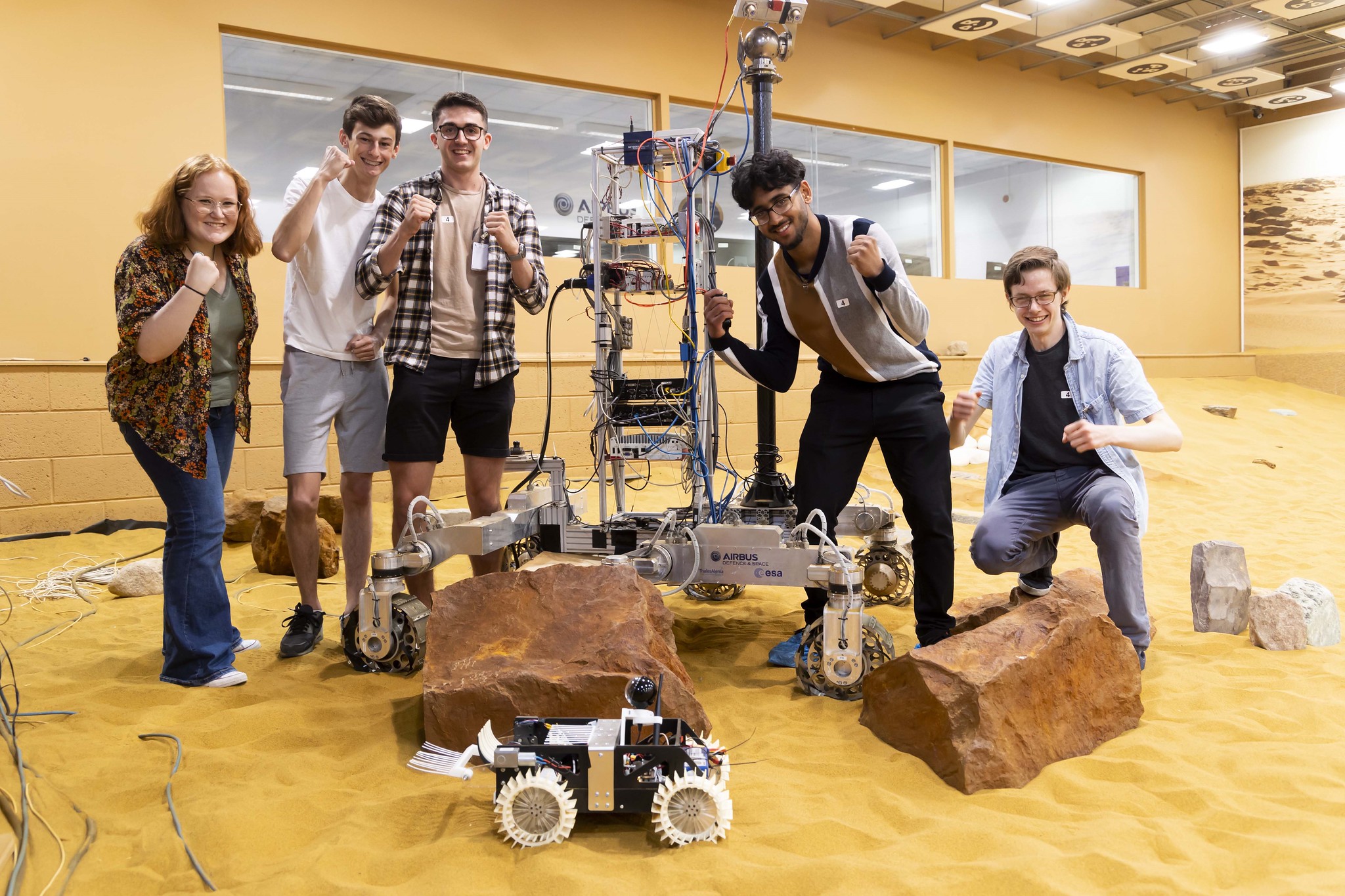 5 people in Airbus's Mars simulation environment. They are standing and crouched around a large robot, with some large rocks and the compeition rover on the floor in front.