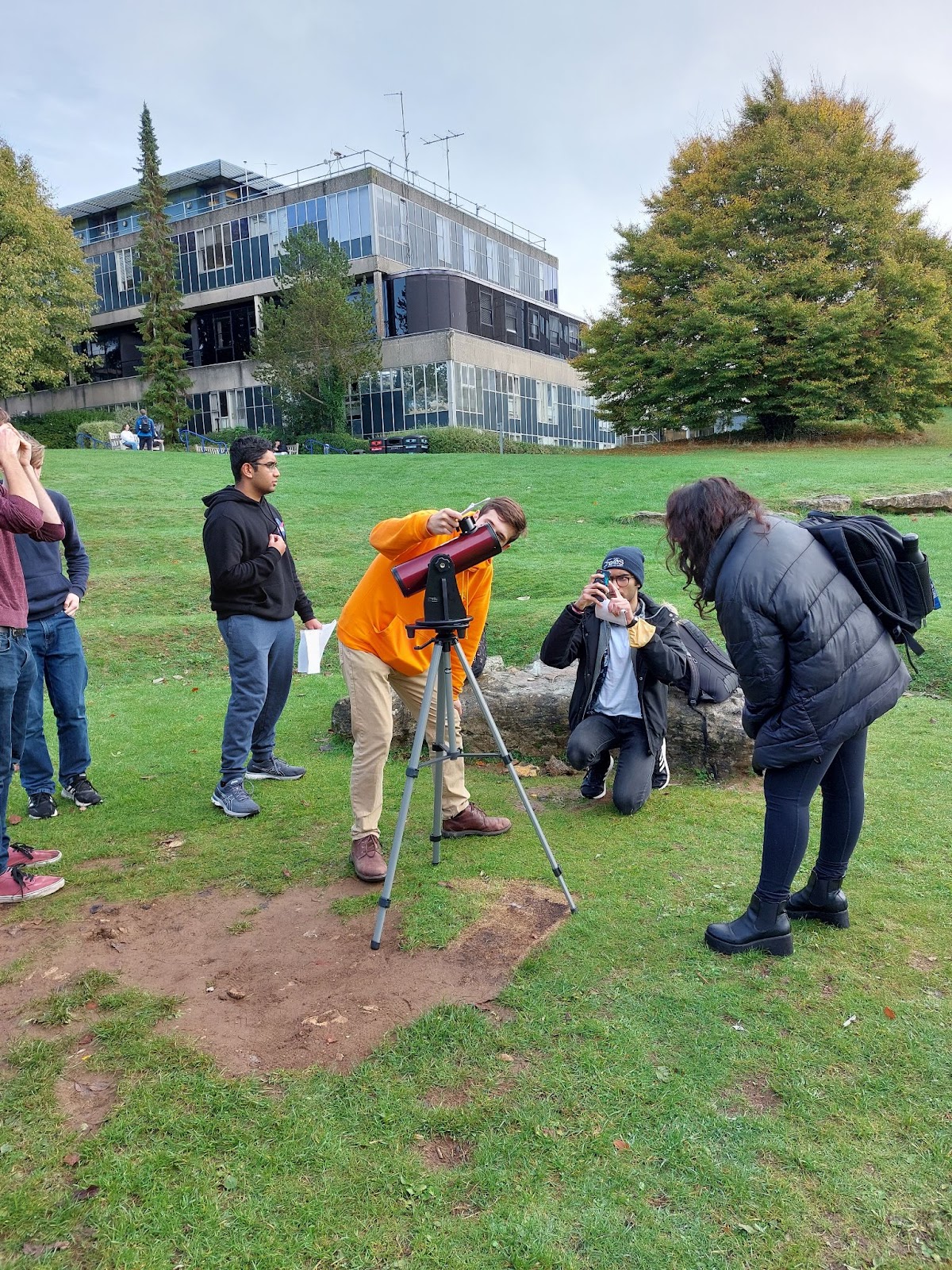 A group of people using a telescope to observe a solar eclipse. A man in orange is creating a projection through the telescope, and various others are standing around