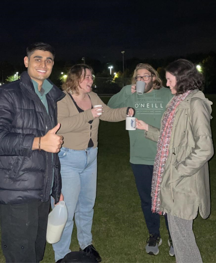 Four people standing outside at night. The leftmost man is grinning with his thumbs up, and the other three people are drinking hot chocolate