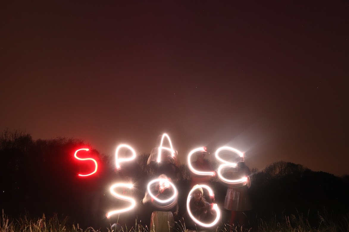 Long exposure art of letters spelling SpaceSoc. A faint glow of aurora can be seen in the background.