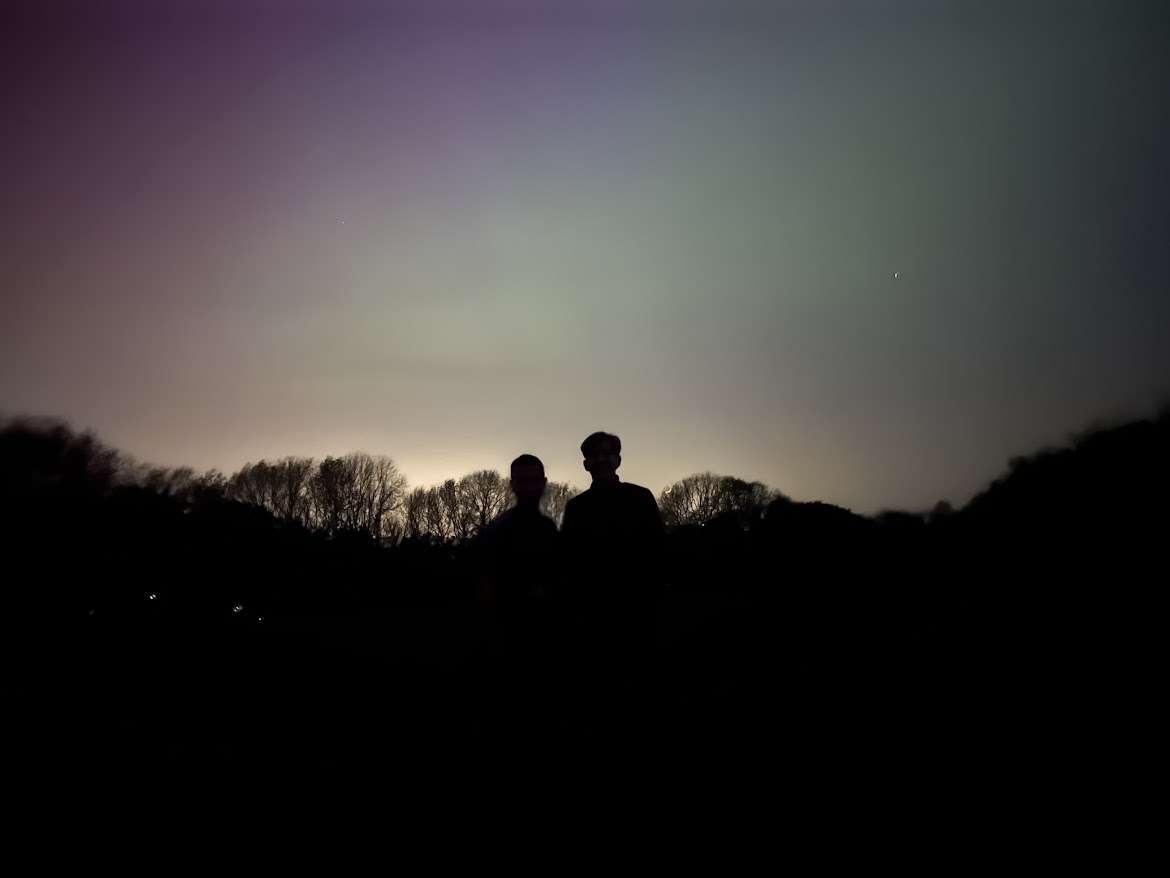 The silhouettes of two students and the treeline contrast the light pollution and aurora in the sky.